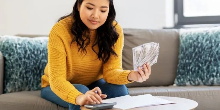 To save money here is an Image of a young girl creating a budget with money in one hand and calculating on a calculator with her other hand