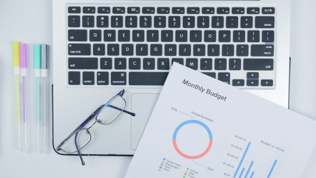 Budget Money for Beginners picture showing a laptop, different color pens, eyeglasses and a piece of paper that says monthly budget showing a budget chart.